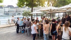 A riverside outdoor seating area with tables and umbrellas, overlooking a river with buildings in the background. - Consum Boutique Hotel (Basel)