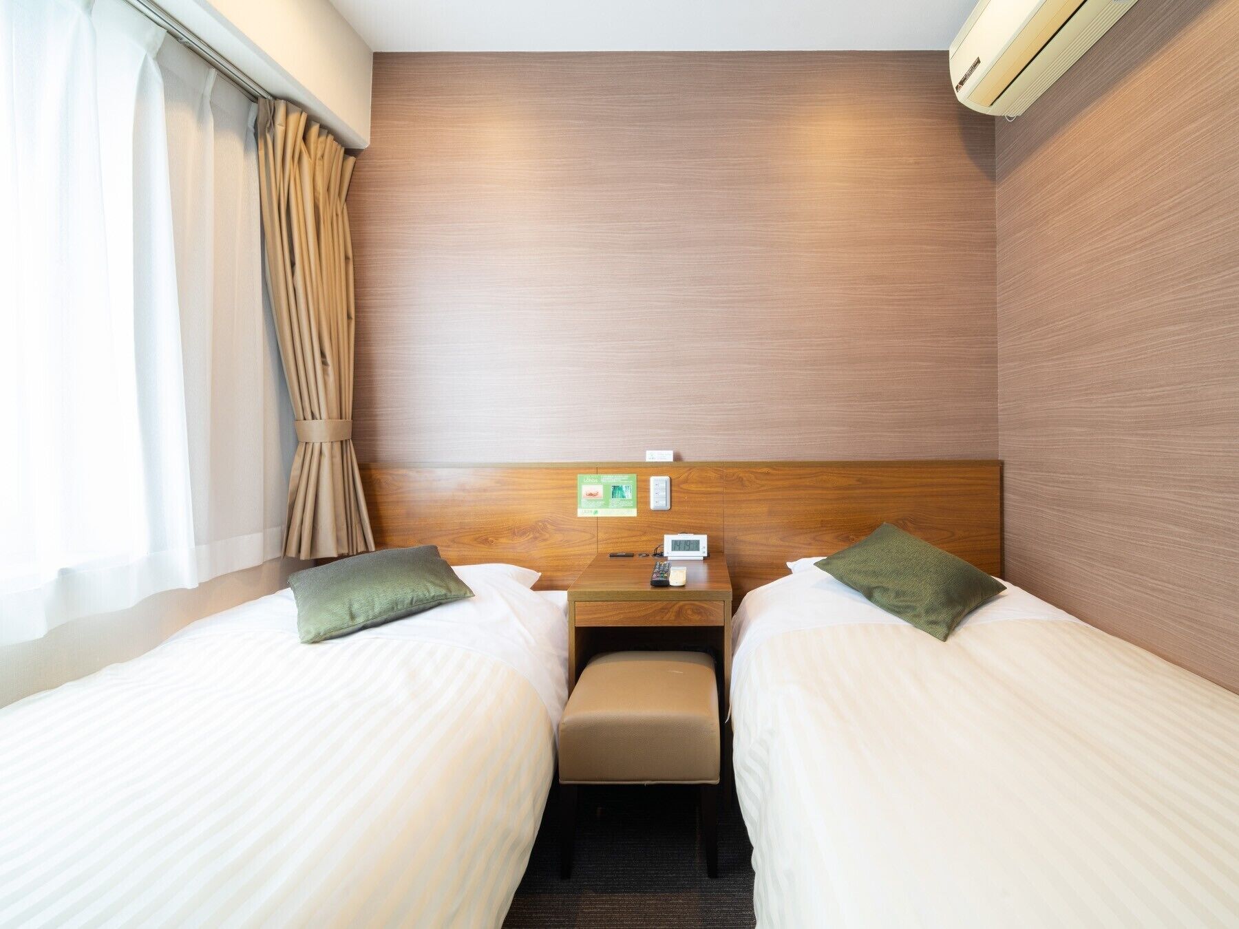 A hotel room with two beds, a small table, a chair, a nightstand, an air conditioning unit, and a window with curtains.