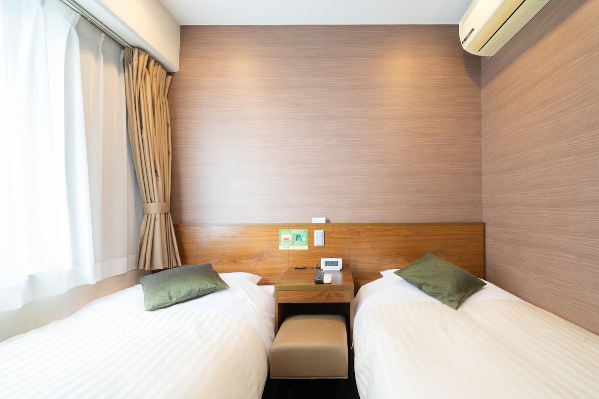 A hotel room with two beds, a small table, a chair, a nightstand, an air conditioning unit, and a window with curtains.