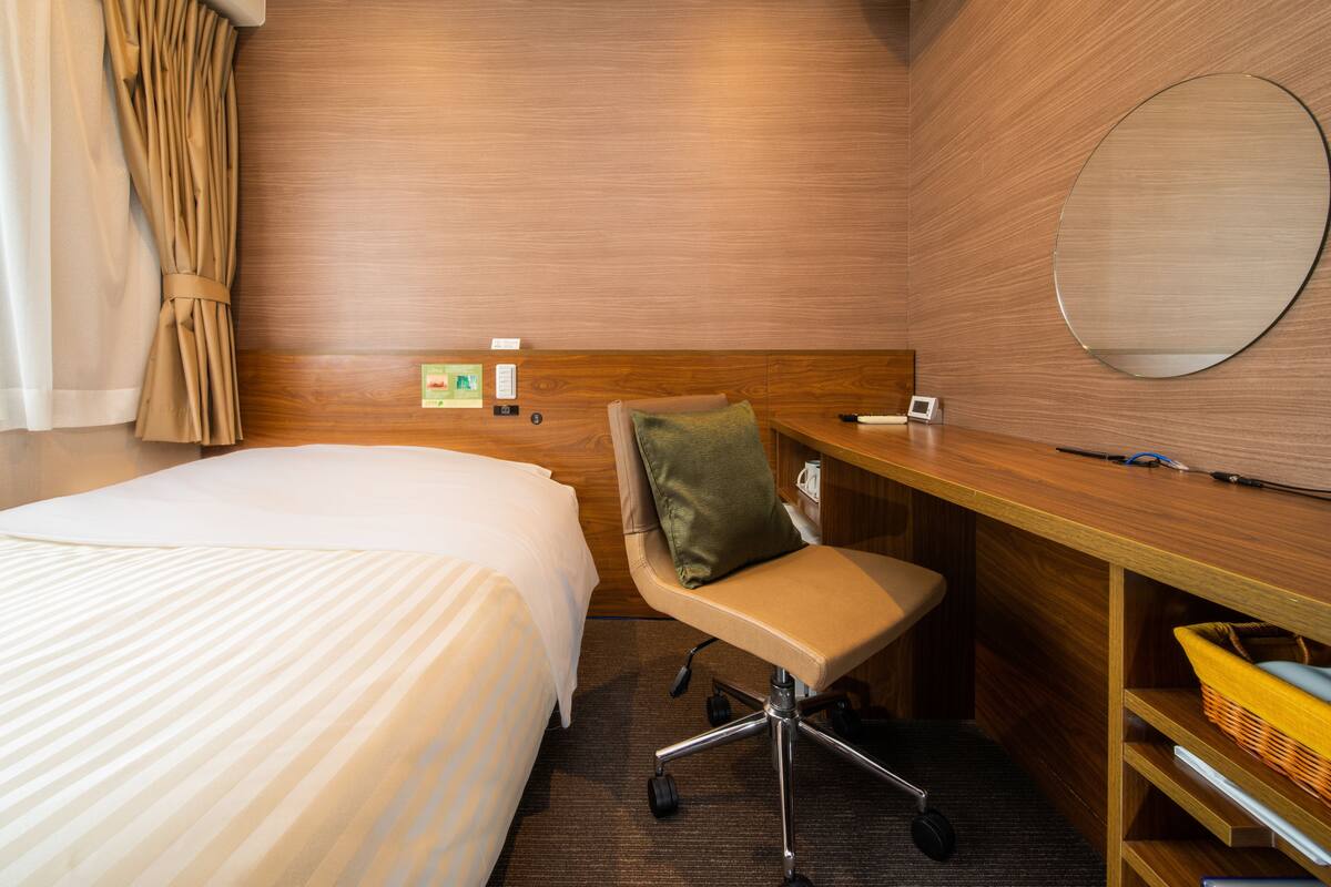 A hotel room with a bed, a desk, a chair, a mirror, and a wall-mounted air conditioner.