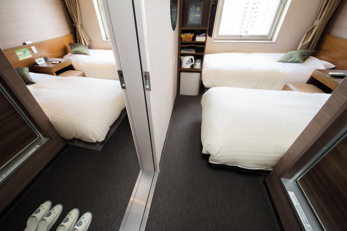 A hotel room with two beds, a small kitchenette, a window, and a shoe rack.