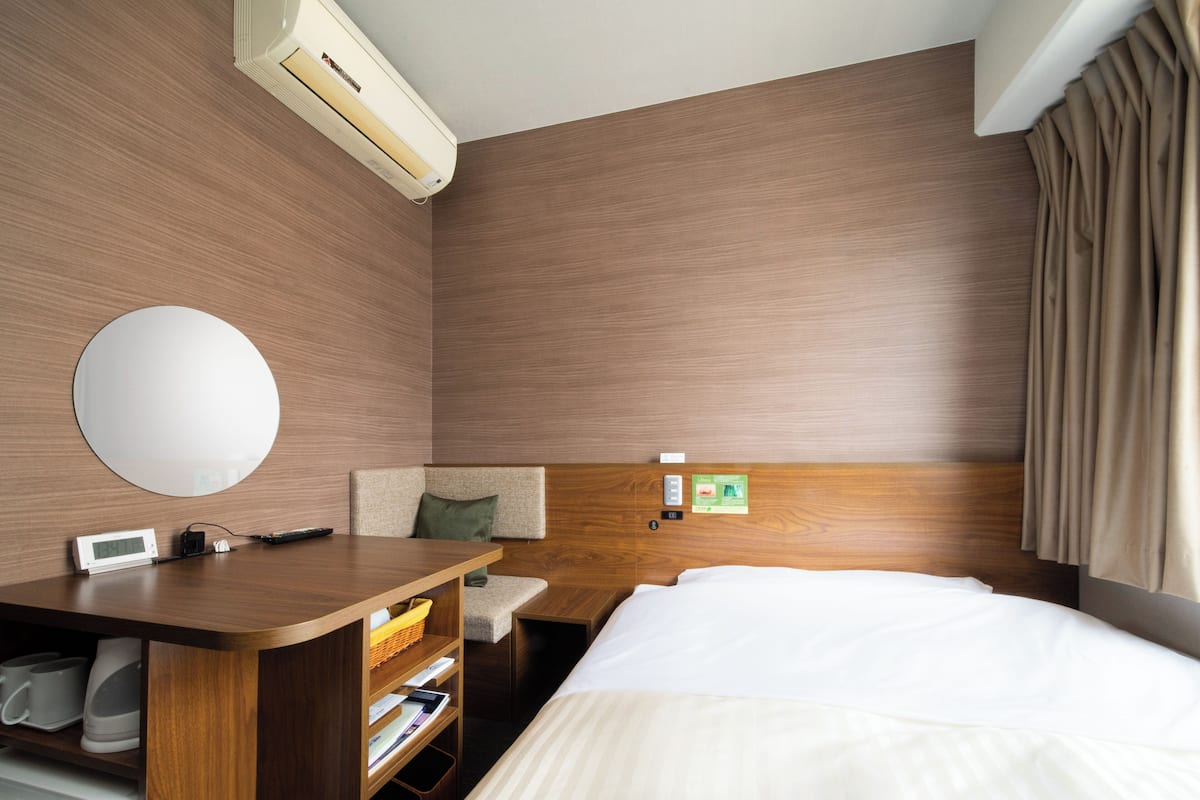 A hotel room with a wooden headboard, a round mirror, a bedside table with a lamp, a bed with white bedding, and a small desk with a basket and papers.