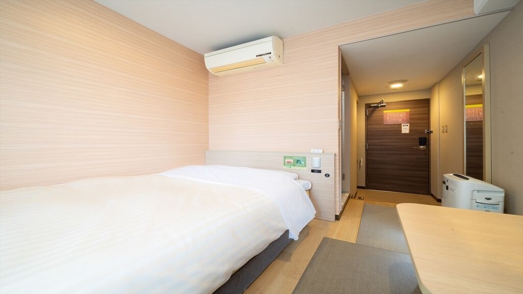 A hotel room with a bed, a desk, a chair, an air conditioning unit, a door, and a trash bin.