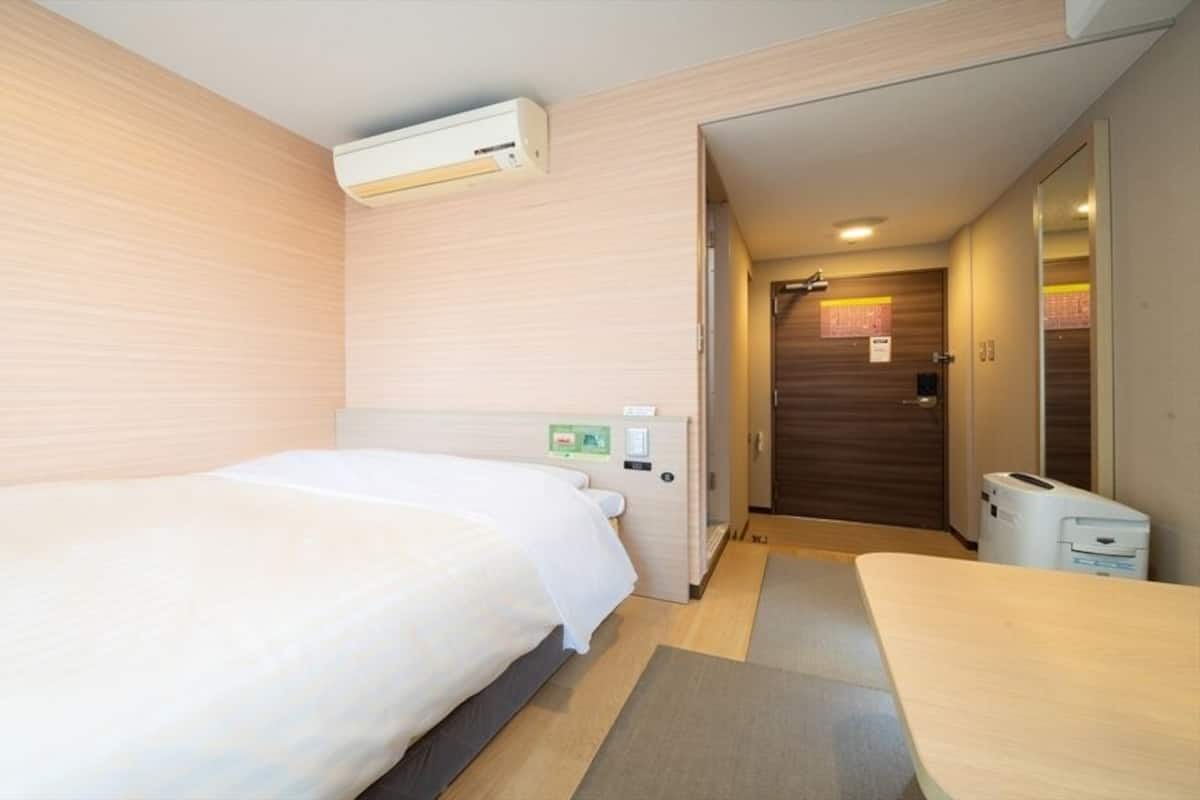 A hotel room with a bed, a desk, a chair, an air conditioning unit, a door, and a trash bin.