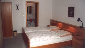 A bedroom with a wooden bed, a bedside table, a nightstand, a mirror, and a small trash bin. - AdiHotels 'Haus Gombert' (Knullwald)