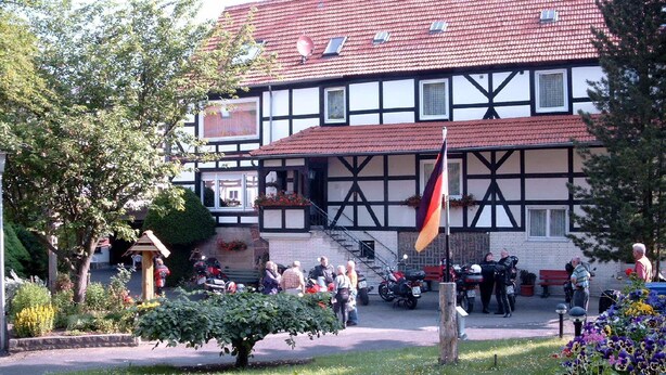 A traditional half-timbered building with a red-tiled roof, a flagpole, and a garden with flowers and a tree. - AdiHotels 'Haus Gombert' (Knullwald)