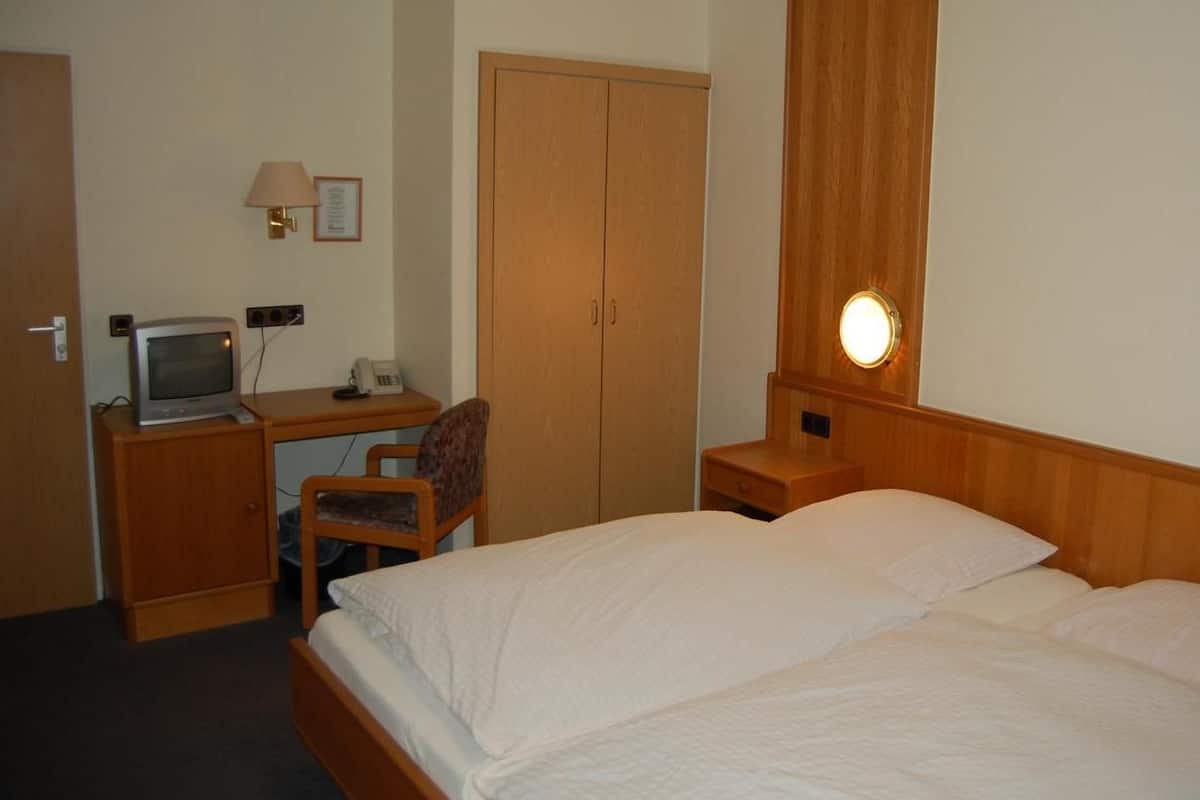 A hotel room with a bed, a desk with a computer, a chair, a wardrobe, and a bedside lamp.