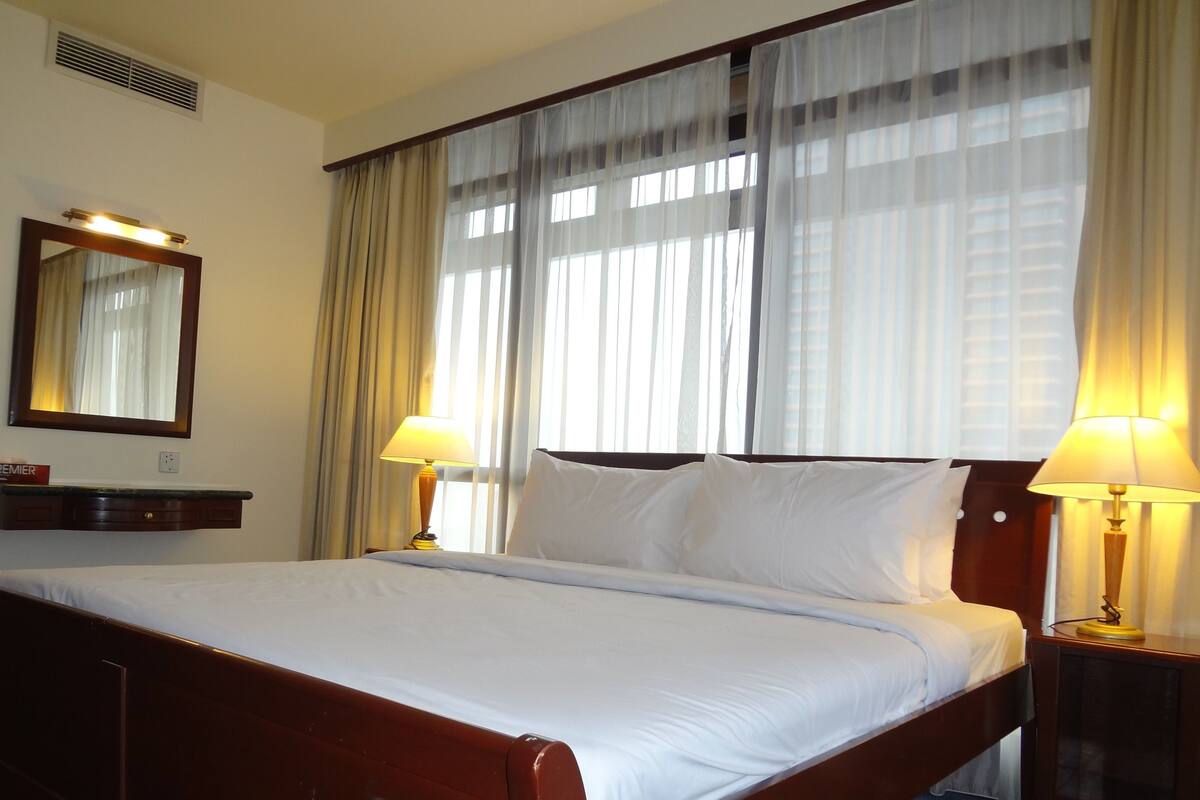 A neatly made bed with white linens, two bedside lamps, a nightstand, a mirror, and sheer curtains on a window.