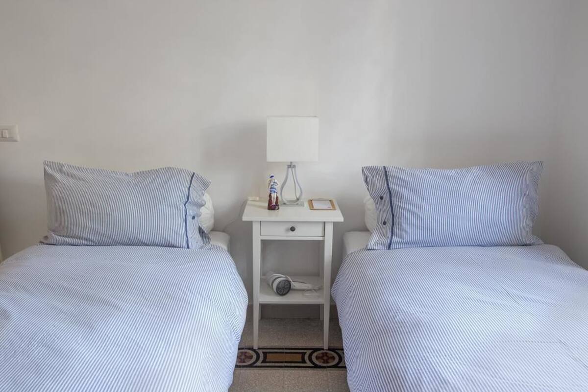 A small, clean room with two single beds, a nightstand with a lamp, and a small shelf.