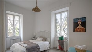 A bedroom with a bed, two windows, a radiator, a bedside table, a hanging light fixture, a painting, and a small table with a basket. - Equilibrì Guest House (Rome)