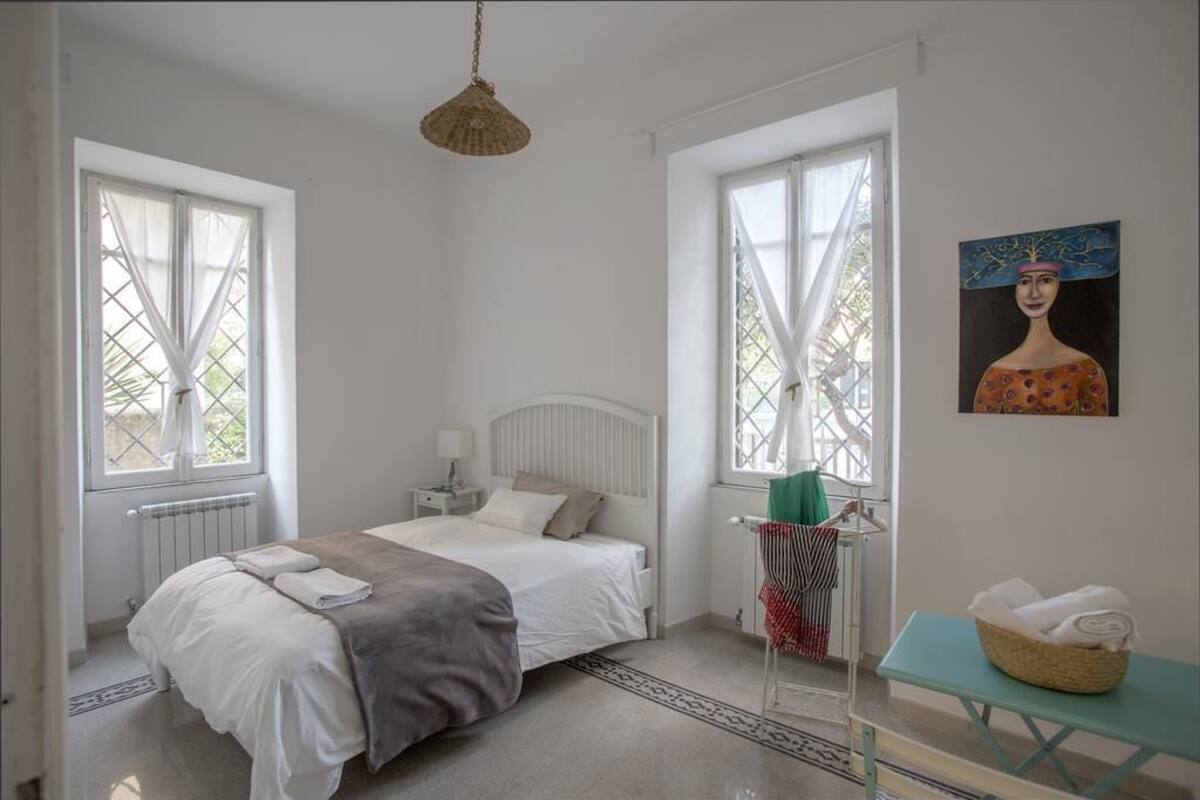 A bedroom with a bed, two windows, a radiator, a bedside table, a hanging light fixture, a painting, and a small table with a basket.