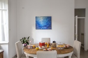 A dining area with a wooden table set for breakfast, a painting on the wall, and a view of the hallway. - Equilibrì Guest House (Rome)