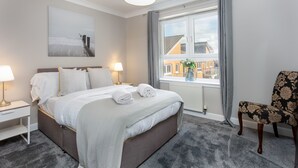 A modern hotel room with a large bed, bedside lamps, a view of buildings through the window, and a chair with a floral pattern. - Harris Retreat - Donnini Apartments (Ayr)