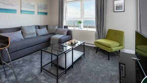 A modern living room with a grey sofa, a glass coffee table, a green armchair, and a view of the sea through a large window. - Harris Retreat - Donnini Apartments (Ayr)