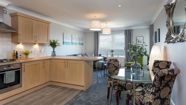 A modern kitchen with wooden cabinets, a central island, and a dining area with floral chairs. - Harris Retreat - Donnini Apartments (Ayr)