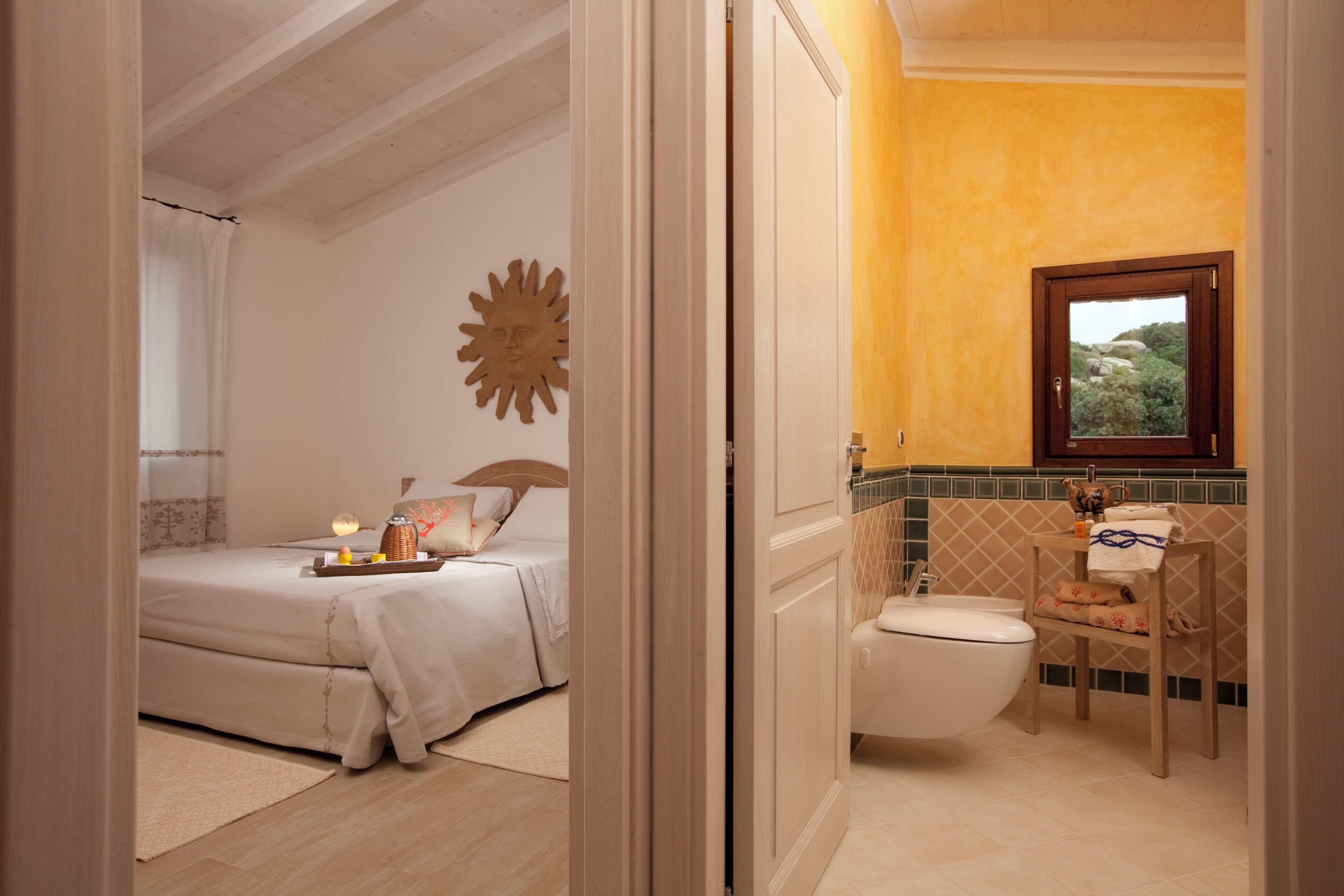 Quarto com cama, mesa de cabeceira e janela. O banheiro tem vaso sanitário, pia e janela.