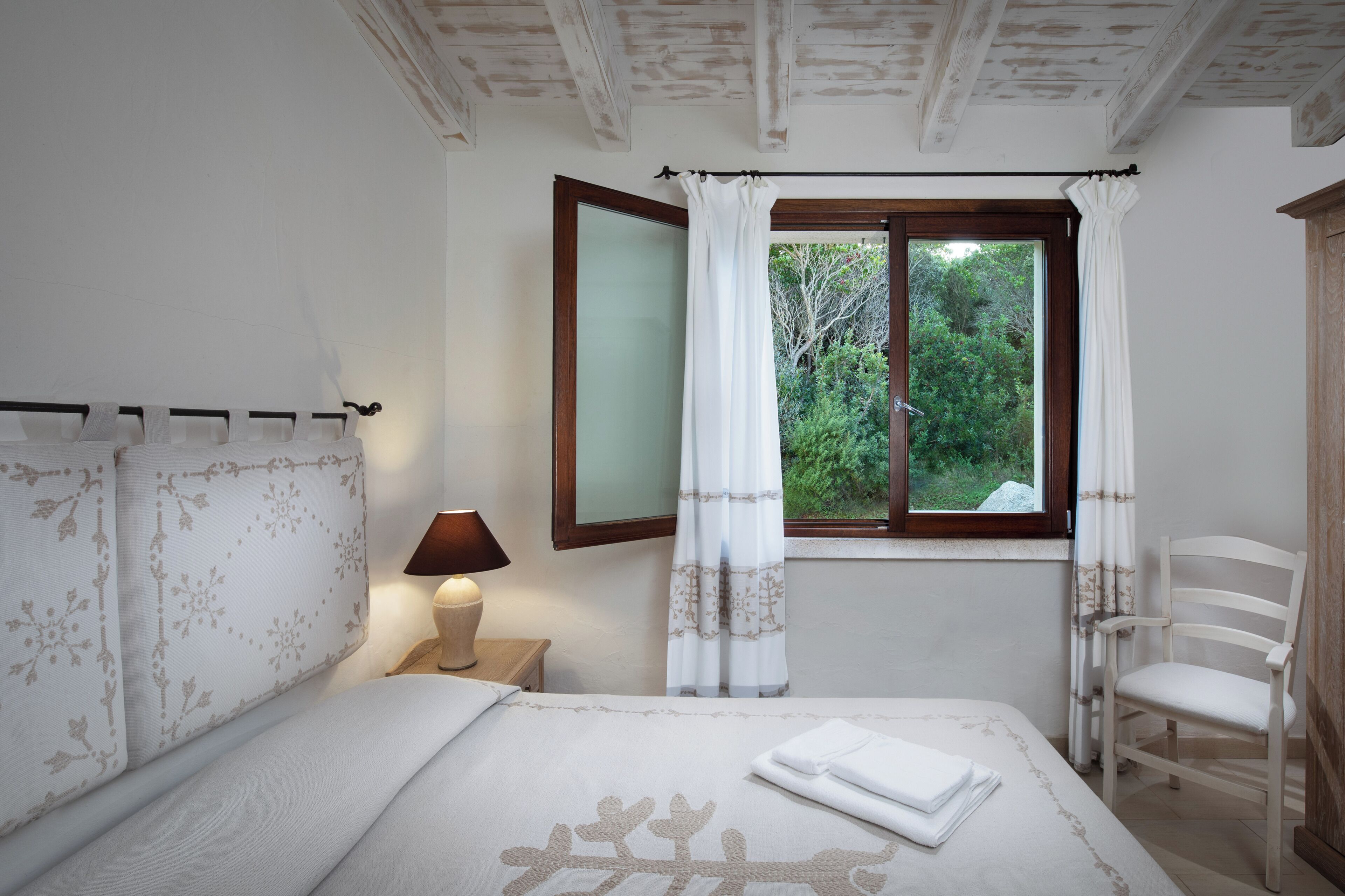 A bedroom with a bed, a nightstand, a lamp, a window with curtains, and a chair.