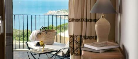 A room with a view of the sea, a table with a basket and fruit, a lamp, and a small wooden side table.
