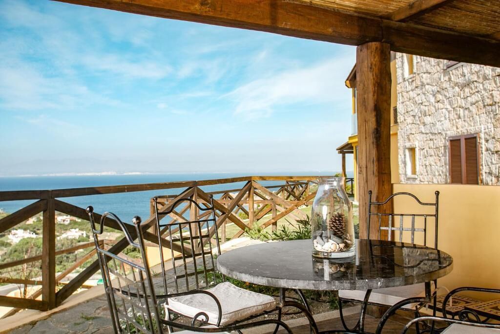 Un espace extérieur aménagé pour les repas, avec une table ronde et des chaises, une structure en verre décorée de pommes de pin et une vue sur la mer.