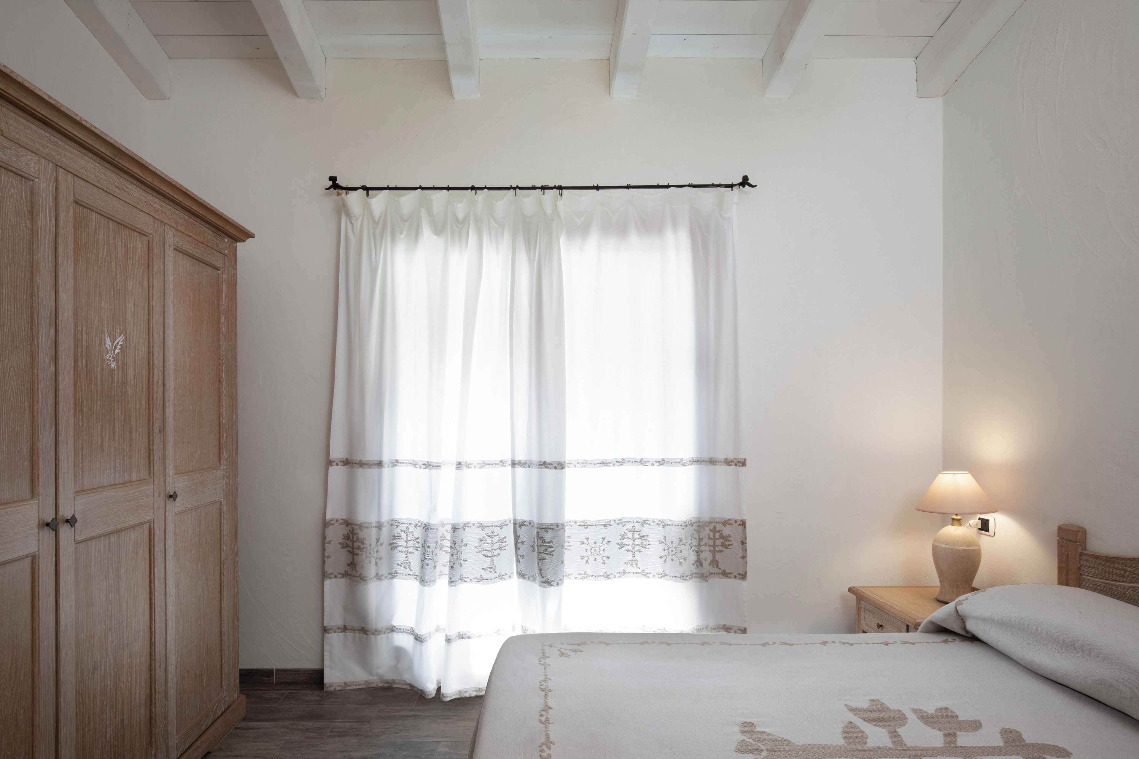 Un dormitorio con un armario de madera, una cama con colcha estampada, una mesita de noche con lámpara y una ventana con cortinas translúcidas.