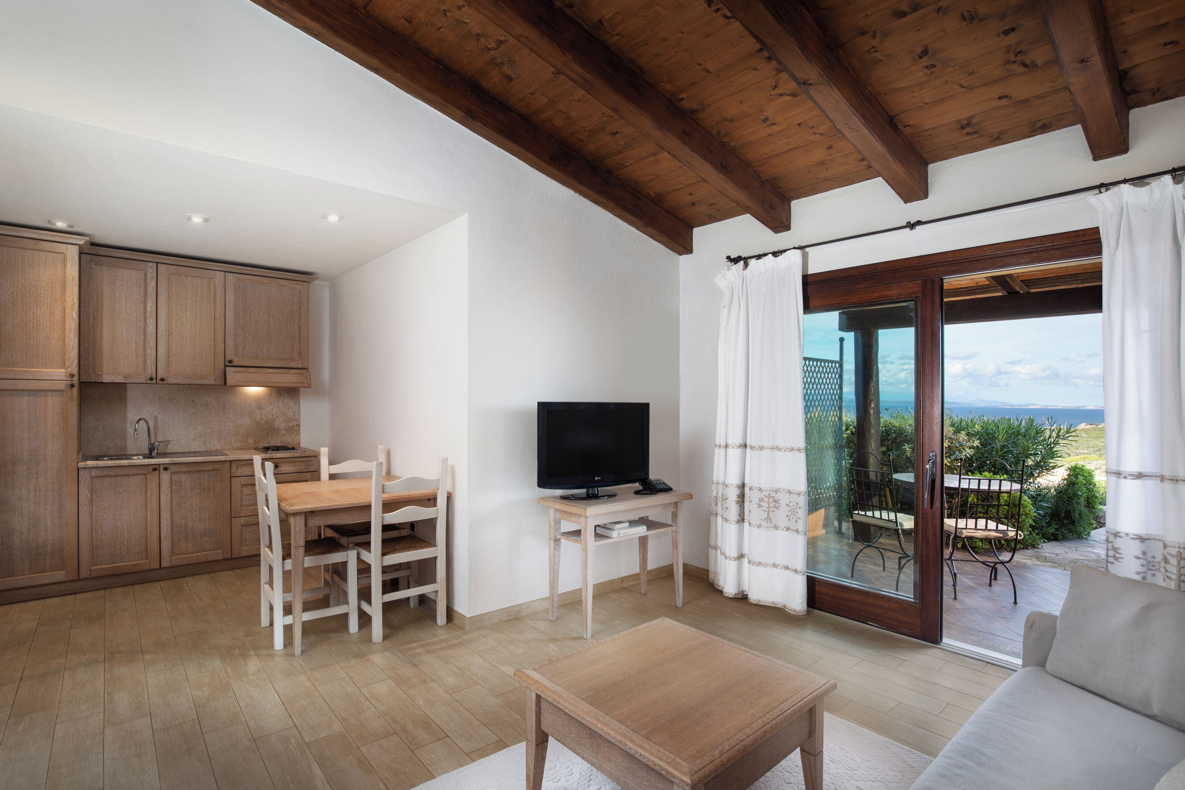 Een moderne woonkamer met een houten salontafel, een bank en een televisie op een bijzettafeltje. Er is een keuken met houten kasten en een eettafel met stoelen. Een schuifdeur leidt naar een terras buiten met uitzicht op het landschap.
