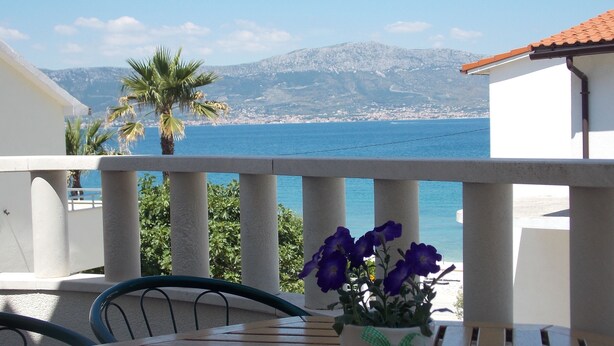 Een balkon met een tafel en stoelen, een bloempot met paarse bloemen en uitzicht op zee en bergen.