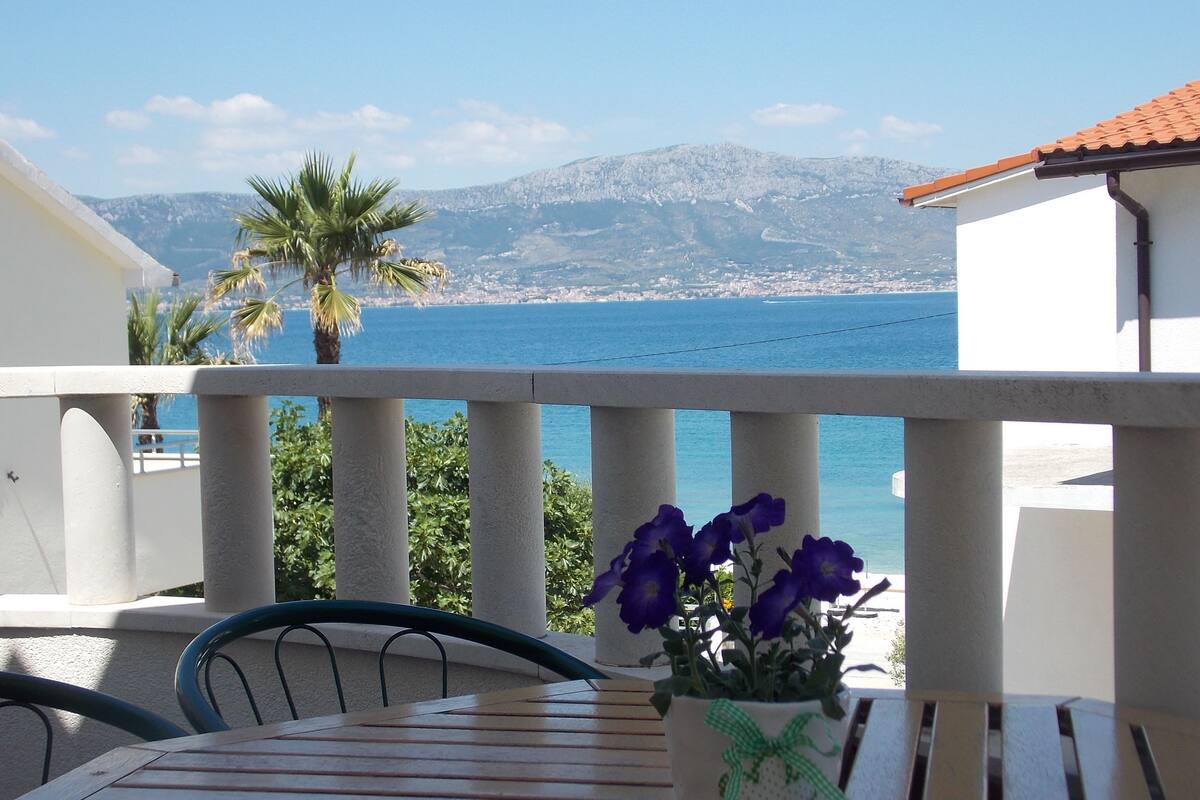 A balcony with a table and chairs, a potted plant with purple flowers, and a view of the sea and mountains.