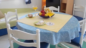 Een eetgedeelte met een ronde tafel, bedekt met een blauwe tafelkleed, omringd door witte stoelen. Op de tafel staat een fruitschaal en een glas sinaasappelsap.