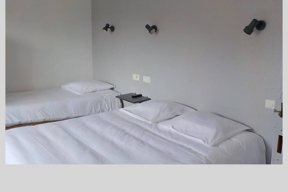 A room with two beds, each with white linens, a small table with a remote control, and wall-mounted light switches.