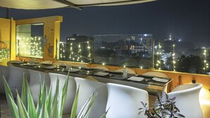 An outdoor dining area with white chairs and a glass-top table, overlooking a cityscape at night. -  Vesta Grand Central Udaipur (Udaipur)