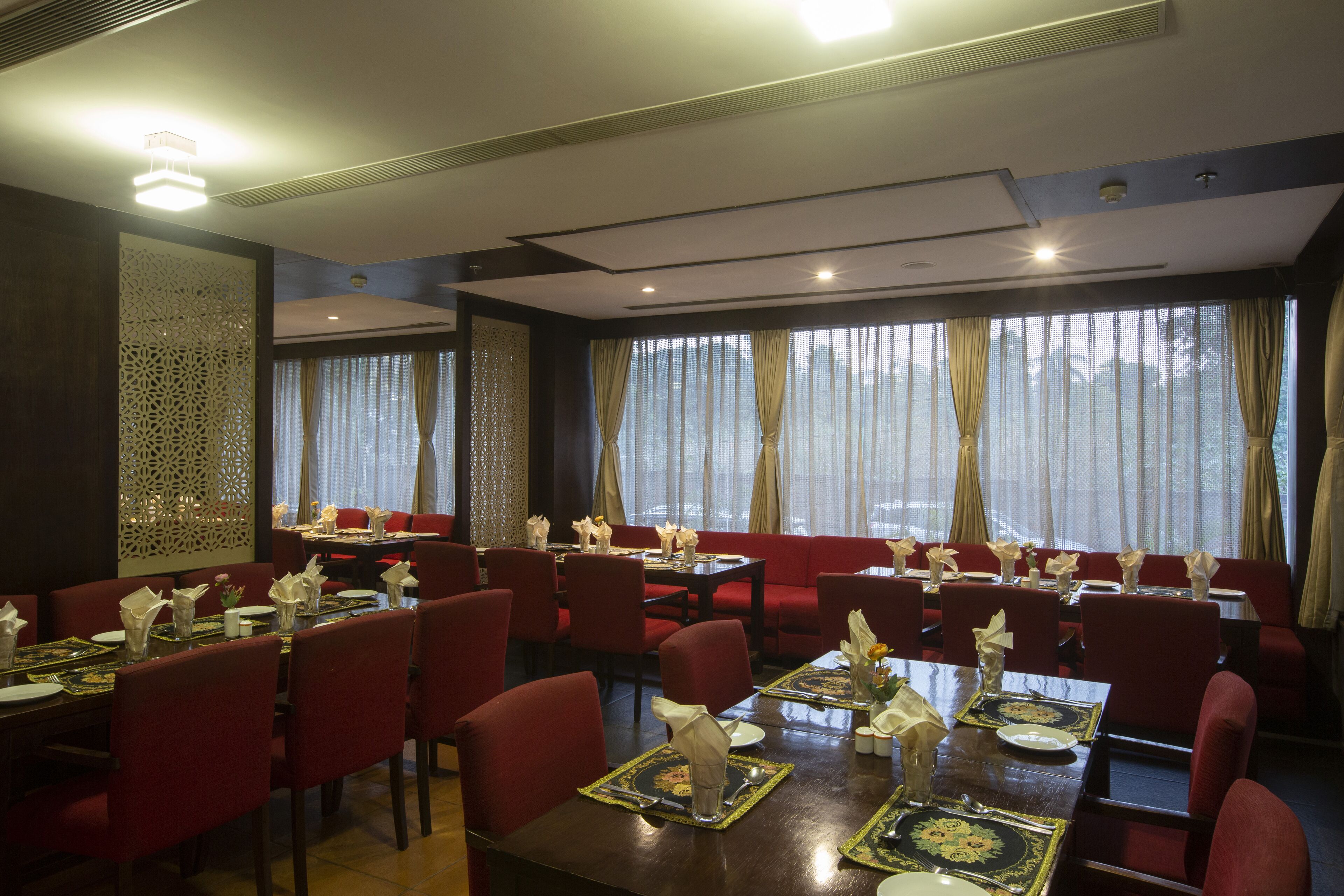 Un restaurante con sillas tapizadas en rojo, mesas dispuestas con manteles blancos y platos, y amplias ventanas con cortinas translúcidas.