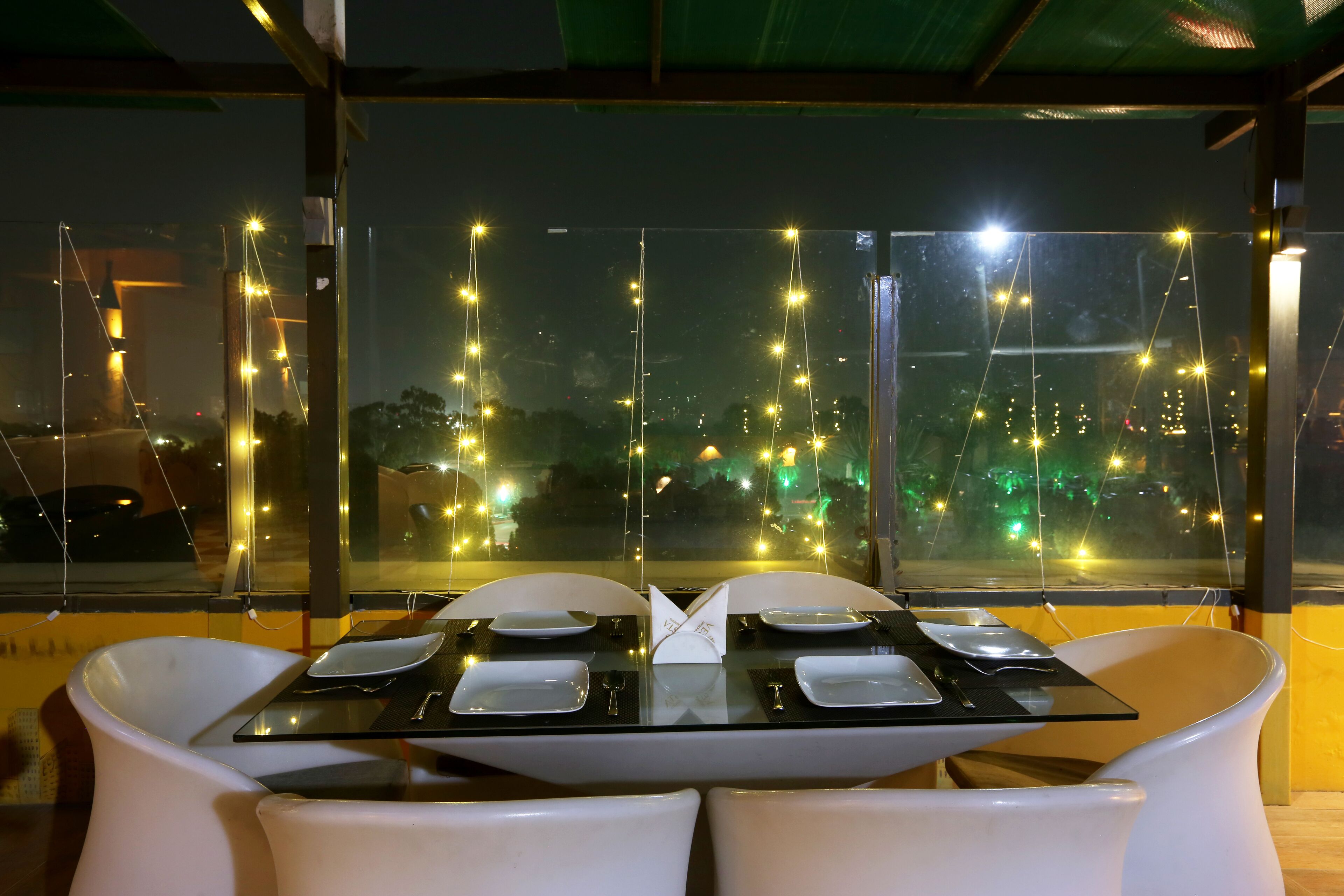 Une table à manger dressée pour deux personnes, avec vue sur le paysage urbain à travers de grandes fenêtres.