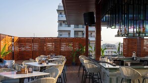 An outdoor dining area with wicker chairs and tables, a bar with a high counter, and a view of the cityscape in the background. -  Vesta Grand Central Udaipur (Udaipur)