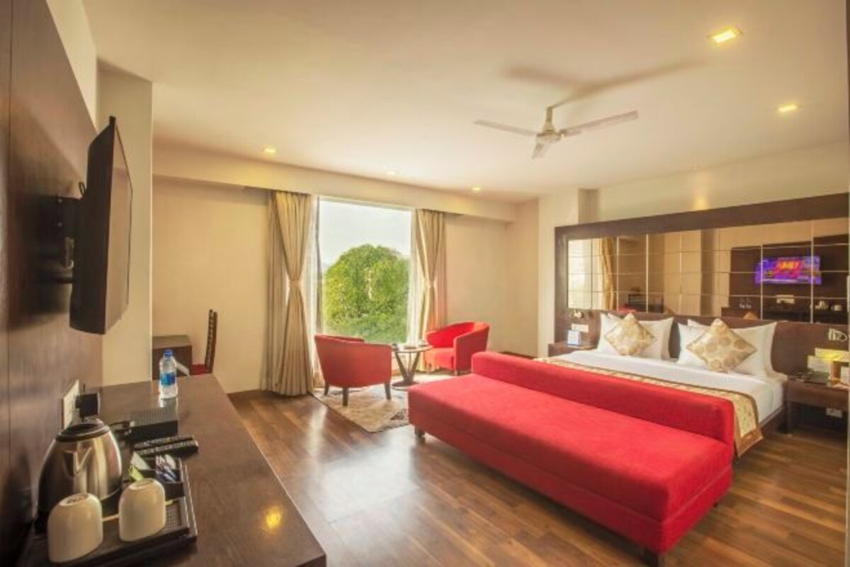 Habitación de hotel con una cama grande, televisión, escritorio con hervidor y tazas, sofá rojo y vistas a zonas verdes.