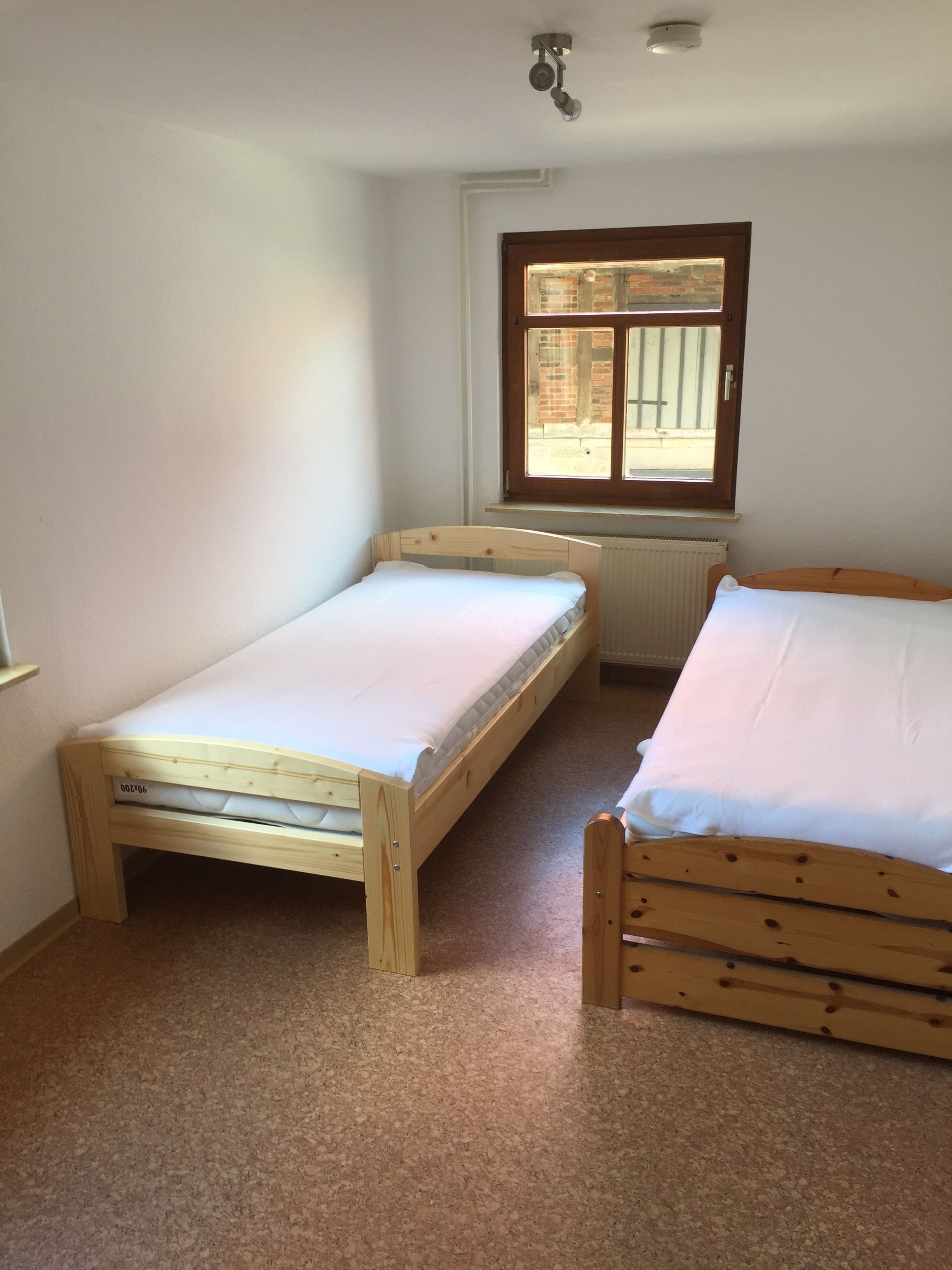Een kleine, nette kamer met twee houten bedden, een raam met uitzicht op een bakstenen gebouw en een plafondlamp.