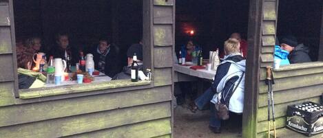 Una cabaña de madera con comedor. Se ven mesas con botellas y vasos, y una mochila en el suelo.