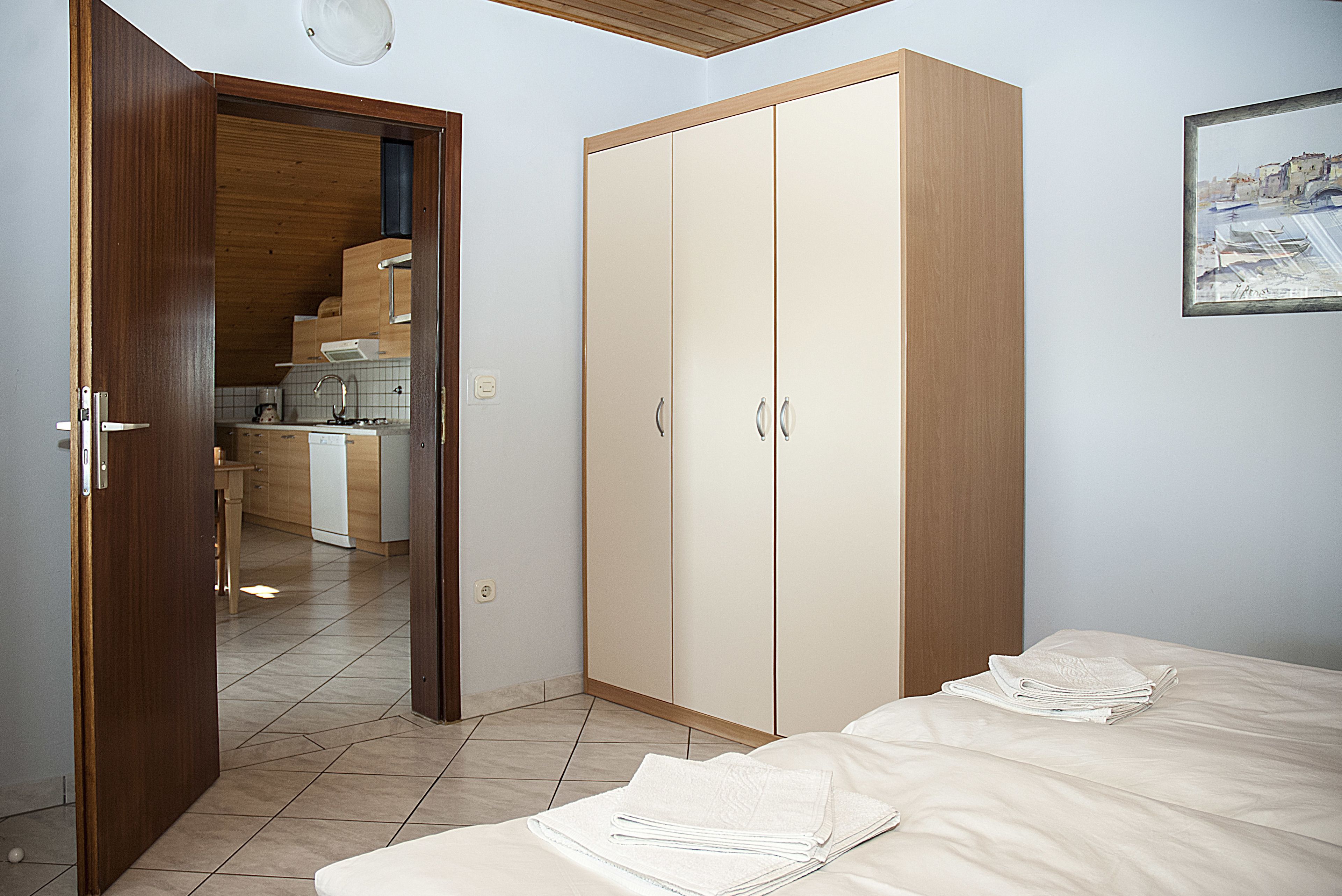 A bedroom with a bed, a wardrobe, and a kitchenette in the background.