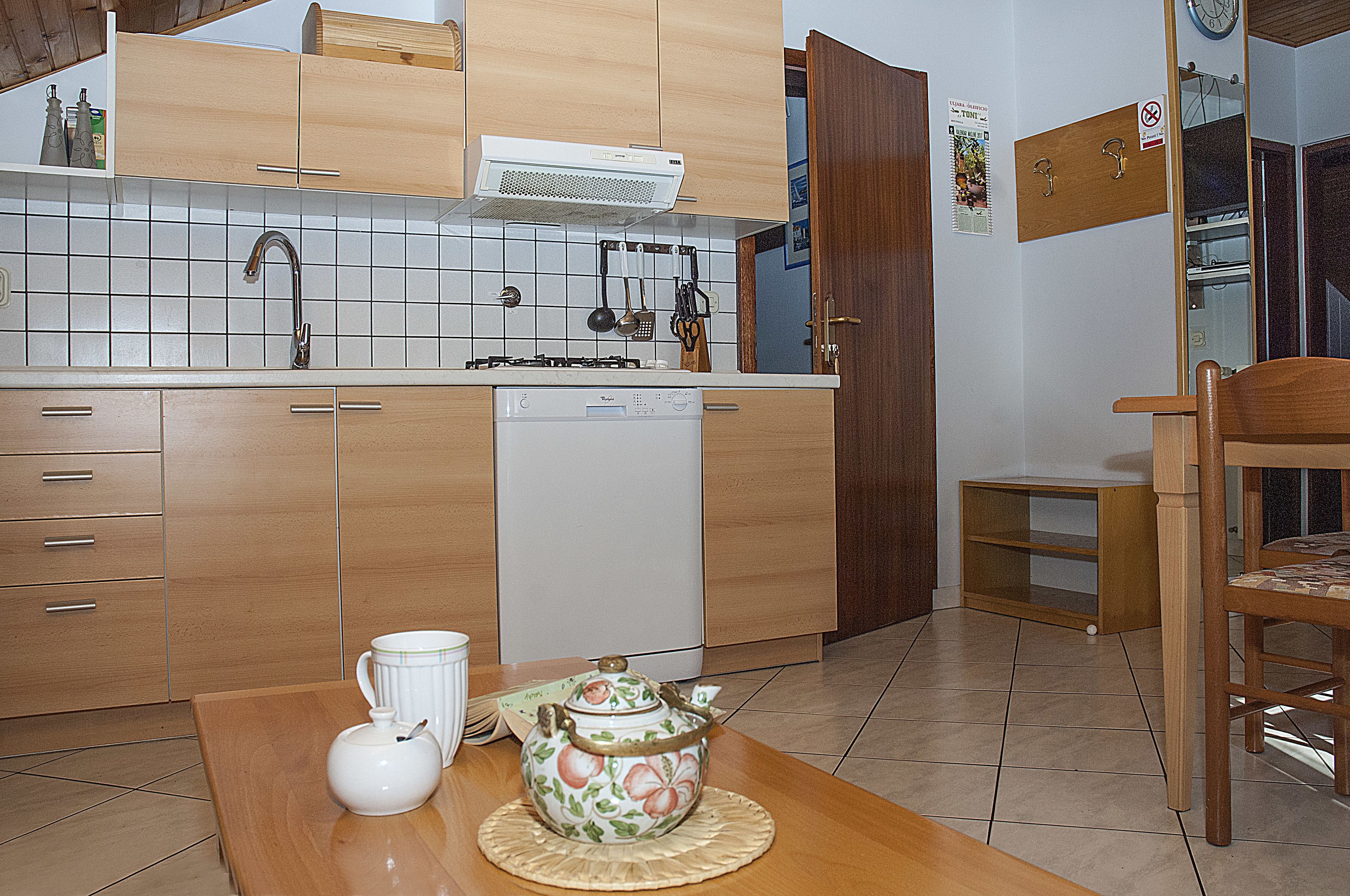 Una cocina compacta con armarios de madera, lavavajillas y una mesa de comedor con tetera y taza.