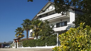 Un edificio de dos pisos con un balcón en el segundo nivel, rodeado de follaje y palmeras.