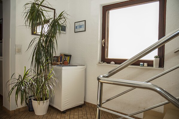 Un escalier propre et bien éclairé, doté d’une rampe métallique, d’une plante en pot, d’une petite étagère avec des objets décoratifs et d’une fenêtre donnant sur l’extérieur.
