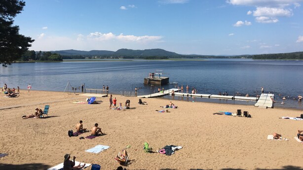 A beach with people sunbathing and swimming, a dock, and a calm lake with mountains in the background. - Ljusdals Camping (Ljusdal)