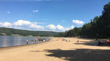 A sandy beach with a calm lake, surrounded by trees and hills.