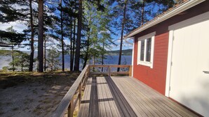 A wooden deck leading to a red building with a white door, overlooking a lake with trees. - Ljusdals Camping (Ljusdal)