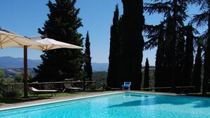 Una piscina azul cristalina con una colchoneta flotando, rodeada de árboles altos y un cielo despejado.