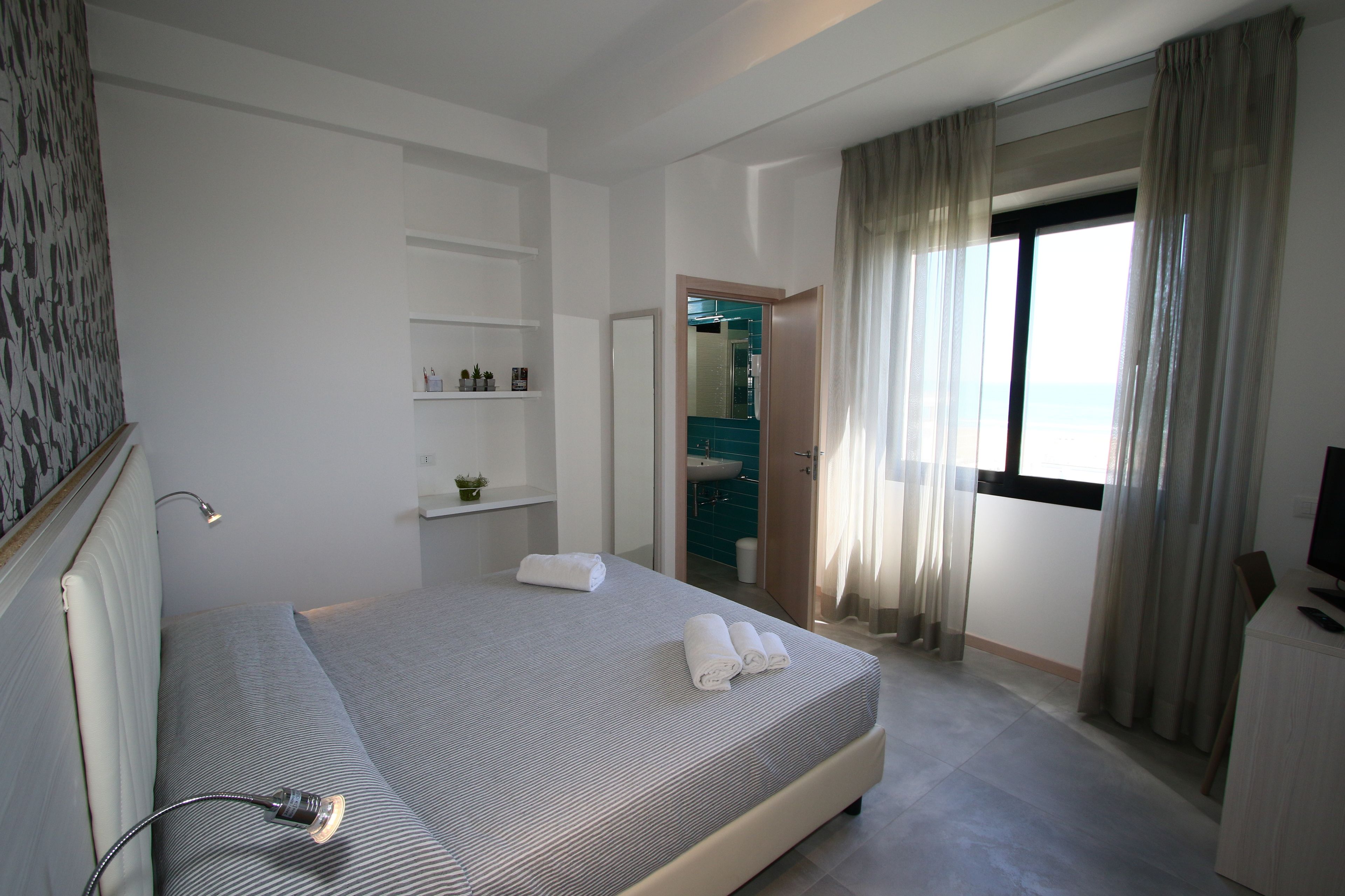 Una habitación de hotel moderna con una cama bien hecha, una lámpara de noche, un pequeño estante con artículos de aseo y vista al mar a través de la ventana.