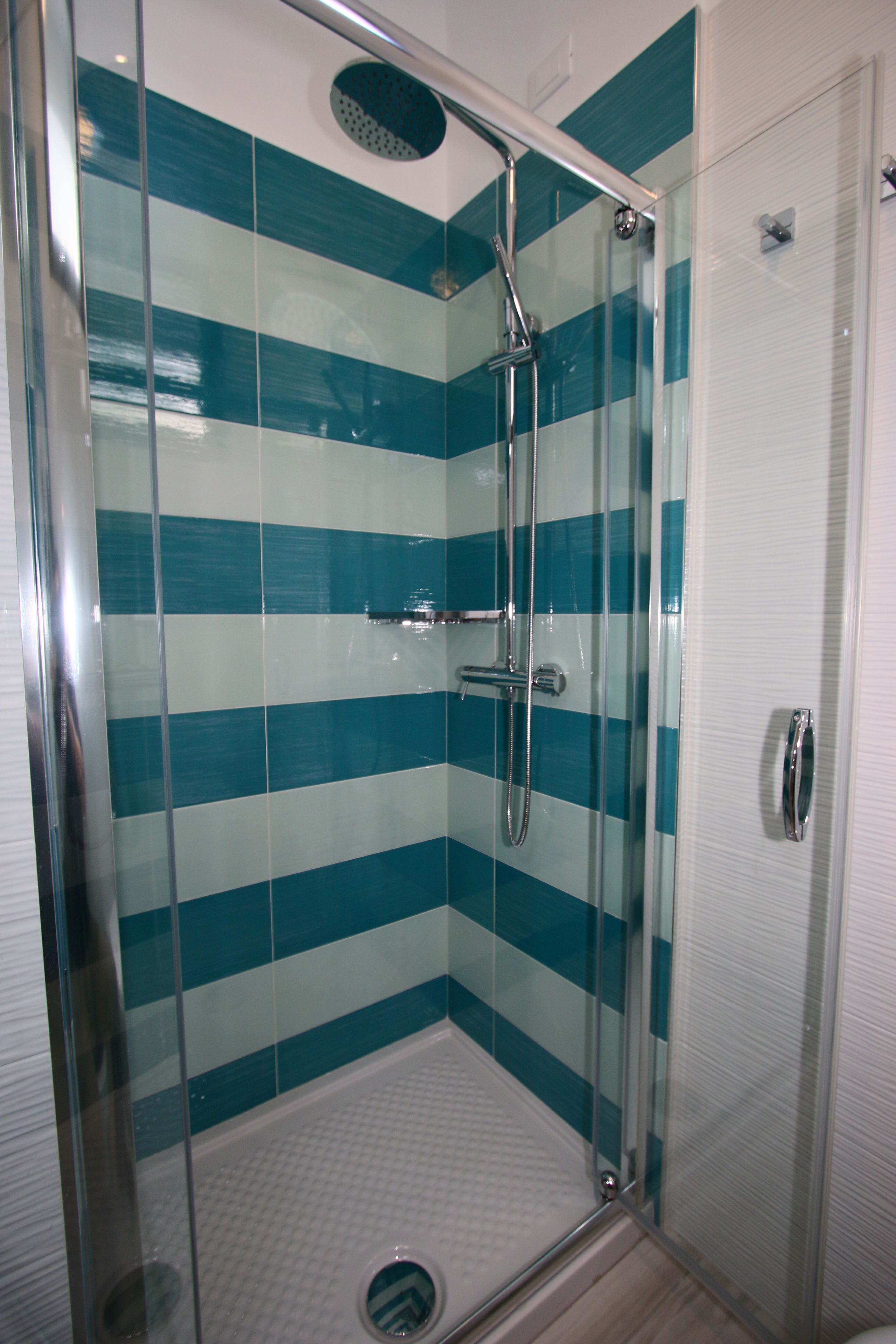 Um chuveiro com divisória de vidro, um chuveiro superior cromado e um chuveiro manual, porta de vidro e piso revestido com azulejos.