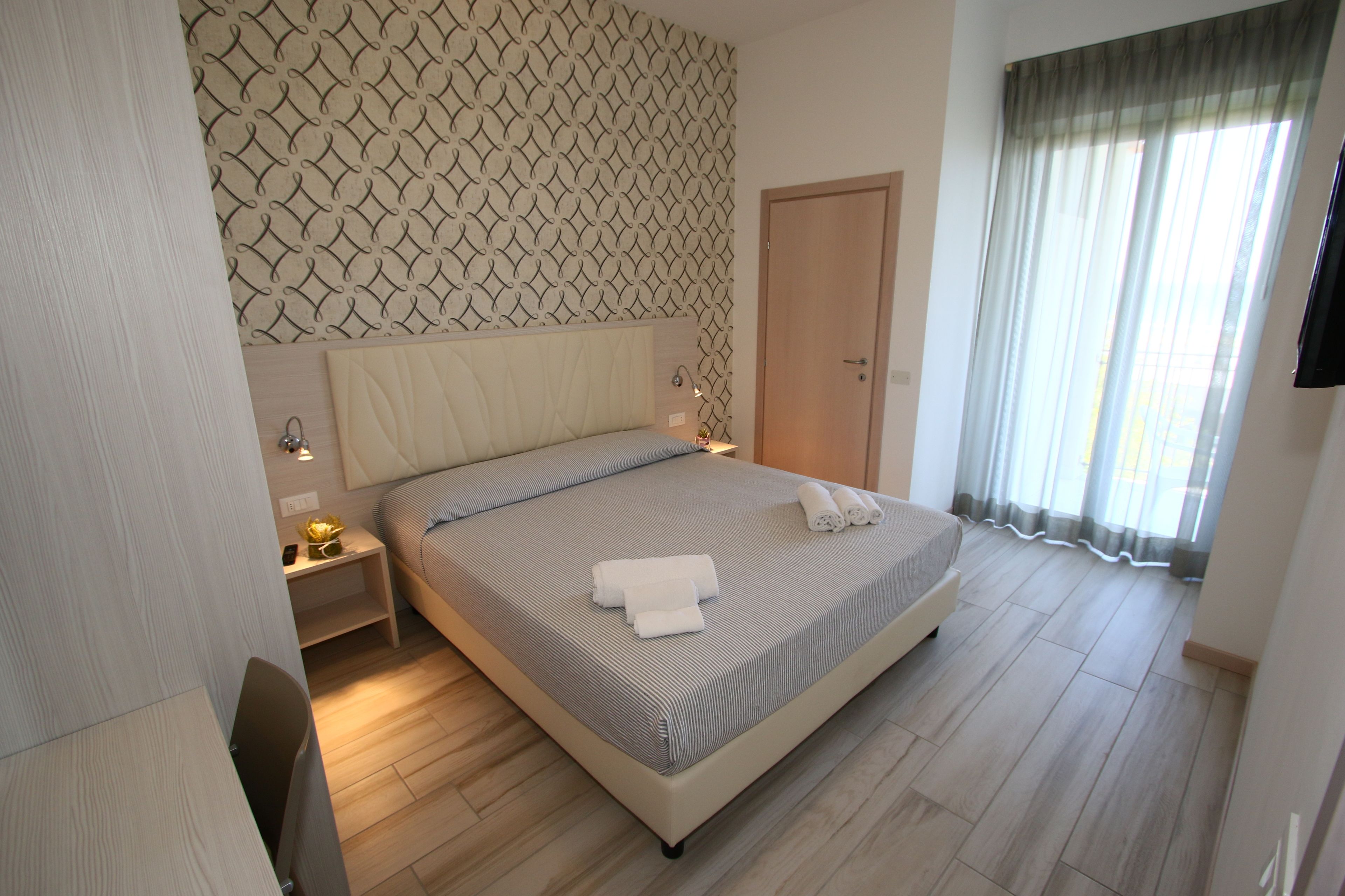 Un letto rifatto con una coperta a righe e due asciugamani arrotolati, un comodino con una lampada, una testiera in legno, un armadio e un'ampia finestra con tende.