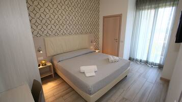 Un letto rifatto con una coperta a righe e due asciugamani arrotolati, un comodino con una lampada, una testiera in legno, un armadio e un'ampia finestra con tende.