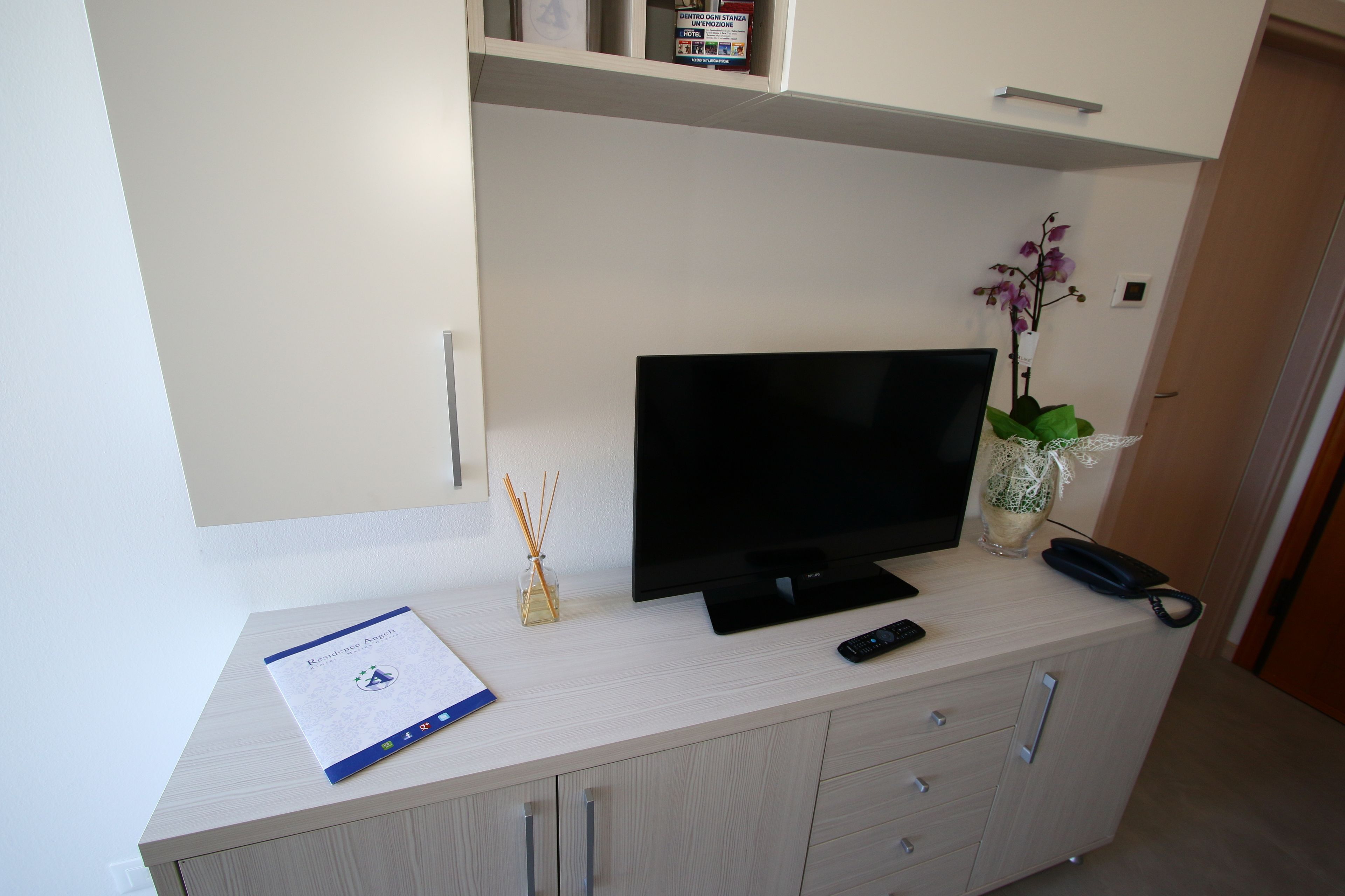 A modern living room with a flat-screen TV on a light-colored cabinet, a remote control, a vase with an orchid, and a small shelf with a box and a candle.