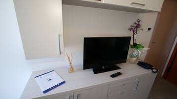 A modern living room with a flat-screen TV on a light-colored cabinet, a remote control, a vase with an orchid, and a small shelf with a box and a candle.